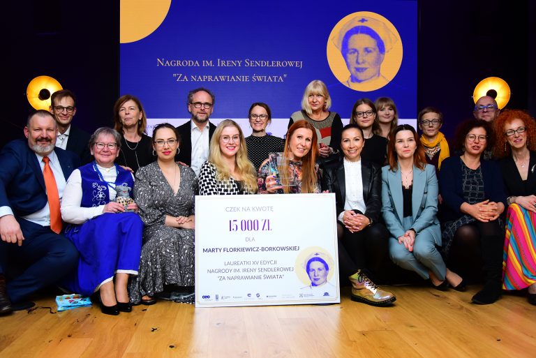 Kobiety i mężczyźni pozują wspólnie do zdjęcia z laureatką Nagrody na tle baneru z wizerunkiem Ireny Sendlerowej. Na dole karta Nagrody z wysokością kwoty 15 tys. zł i nazwiskiem laureatki.